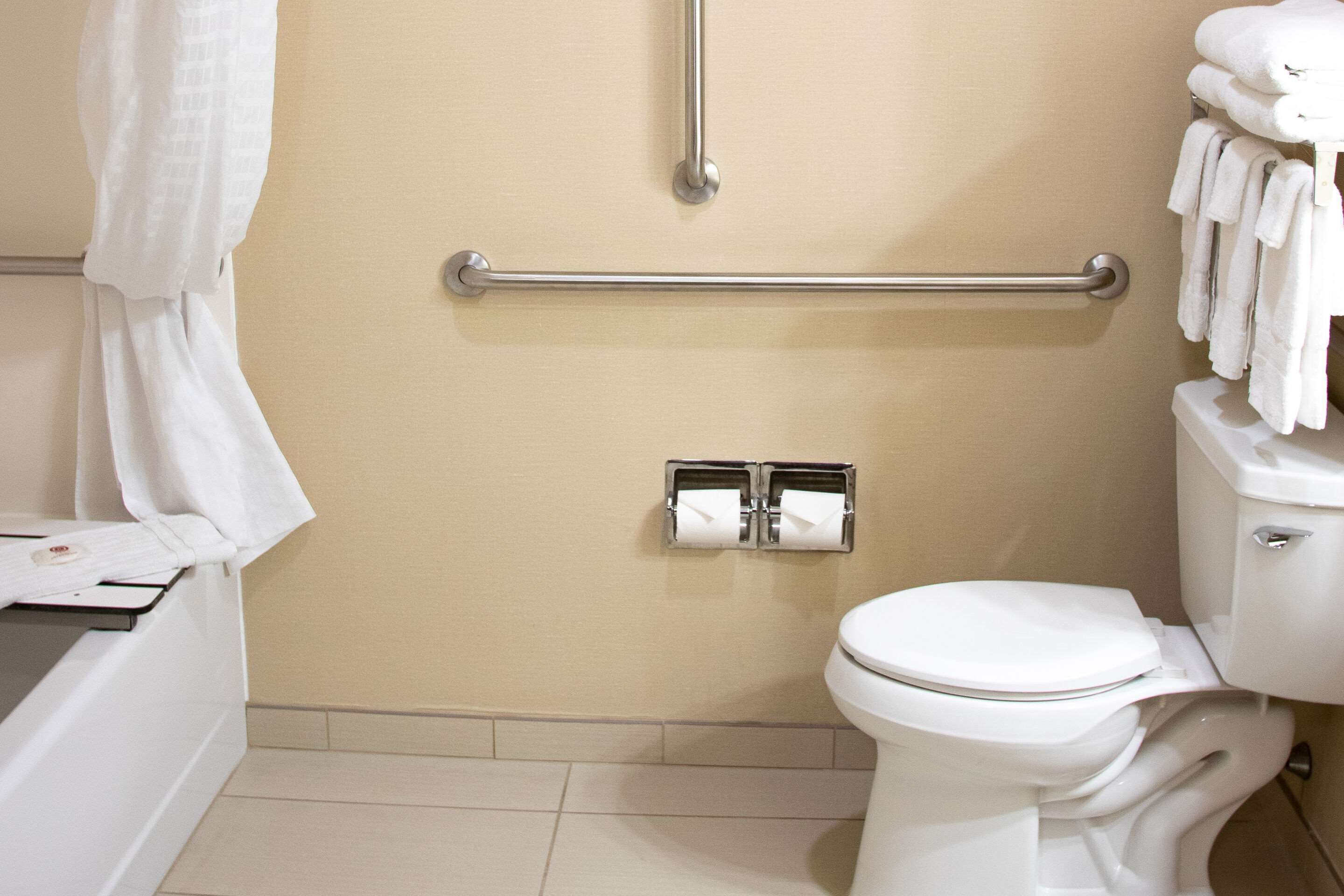 A bathroom with a toilet, a towel rack with towels, a handrail, and a paper towel dispenser.