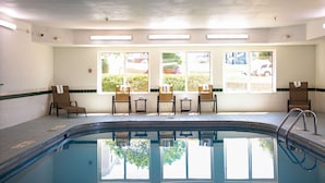 A swimming pool with a sunlit interior, a row of chairs, and a view of the outside through large windows.