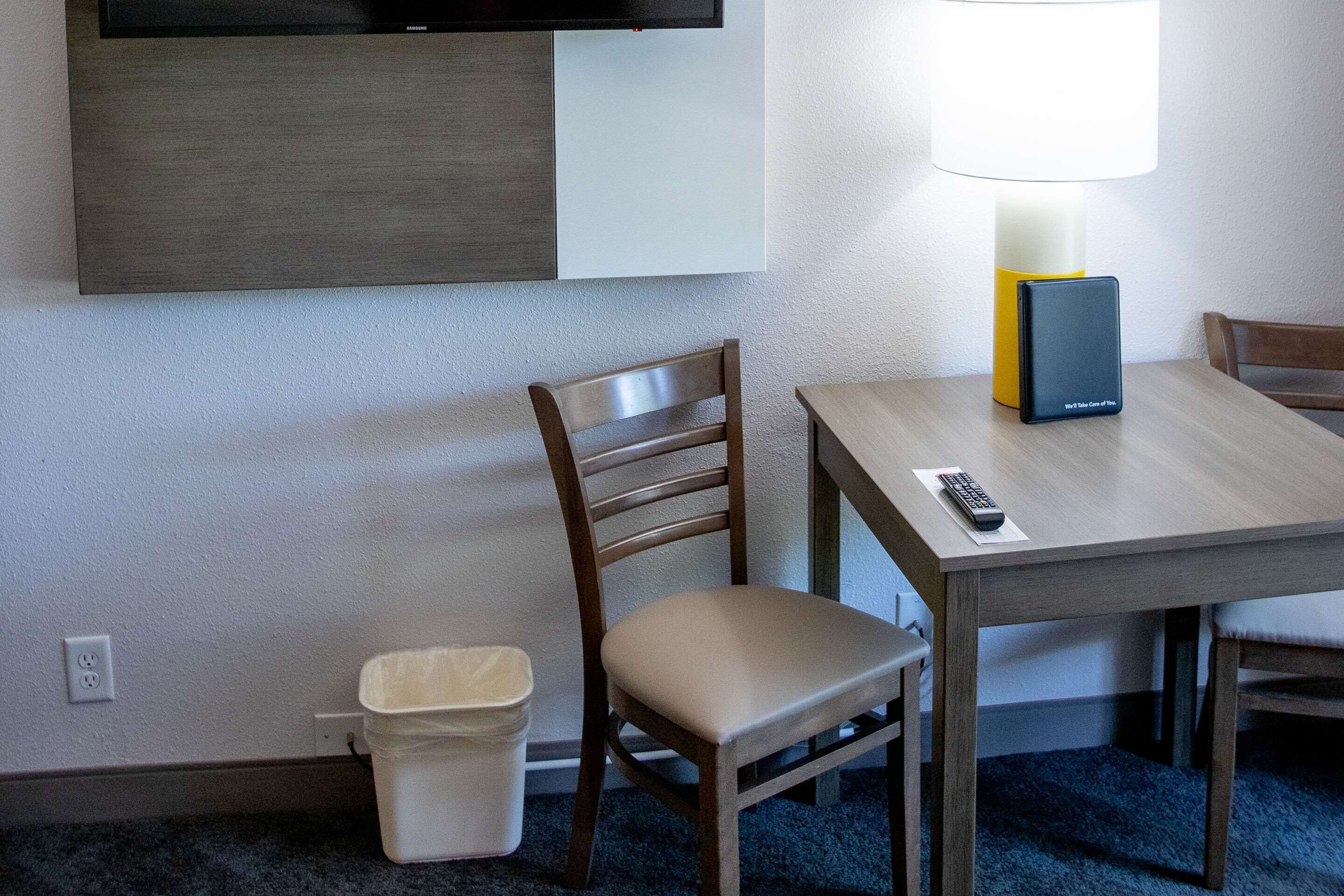 Habitación de hotel con una mesa de madera, una silla, una lámpara, un cubo de basura y un televisor montado en la pared.
