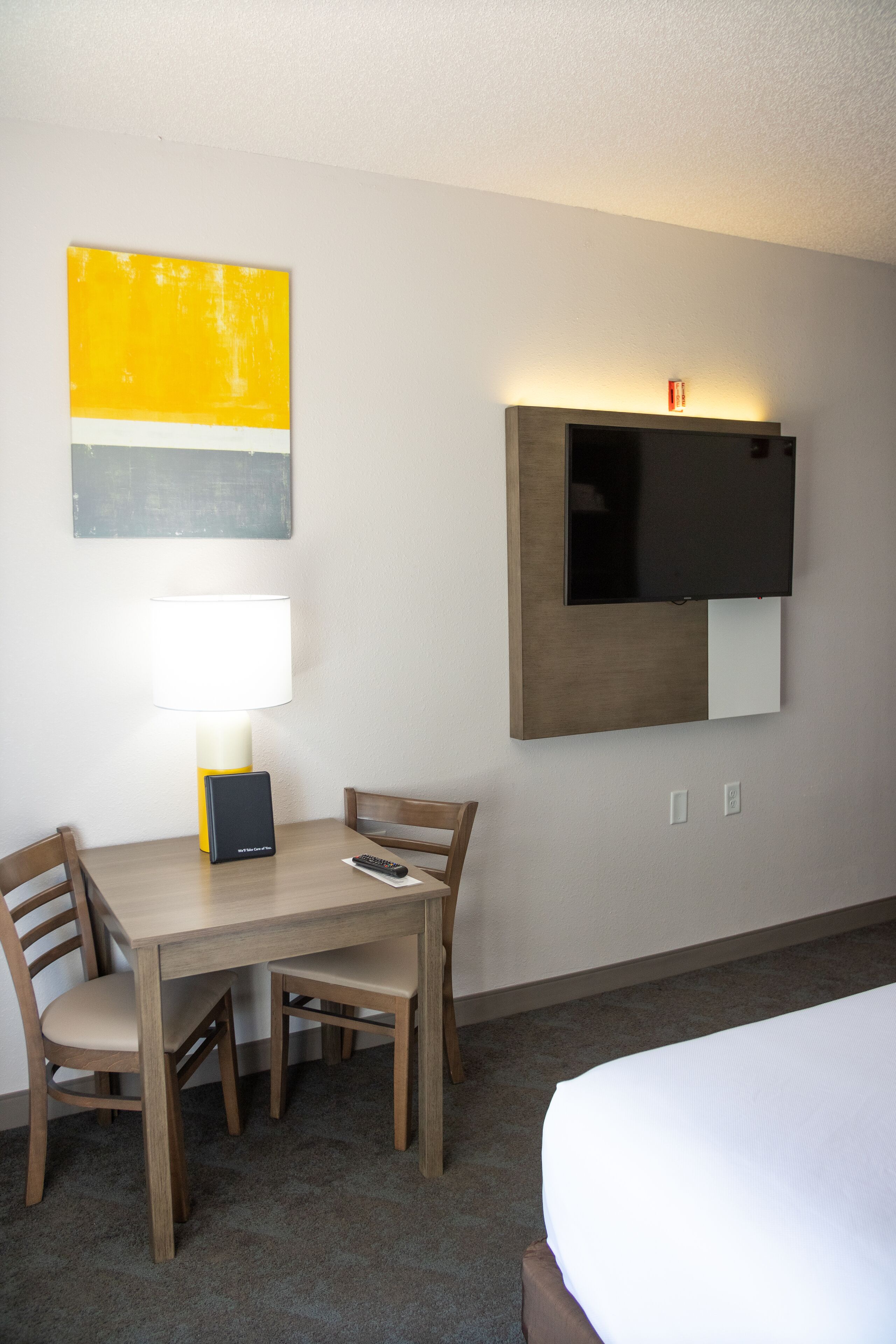 A hotel room with a wooden table, two chairs, a bed, a TV mounted on the wall, a lamp, and a painting.