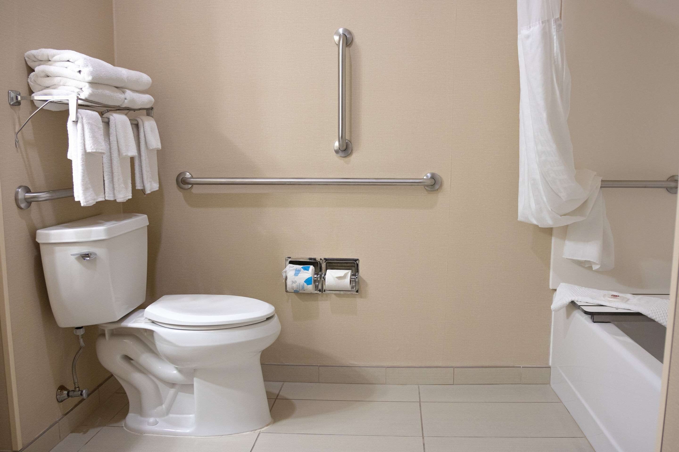 Un baño con inodoro, un perchero con toallas, barra de apoyo y dispensador de papel higiénico.