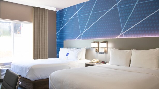 A hotel room with two beds, a desk, a chair, a nightstand with a phone, and a wall with a geometric pattern.
