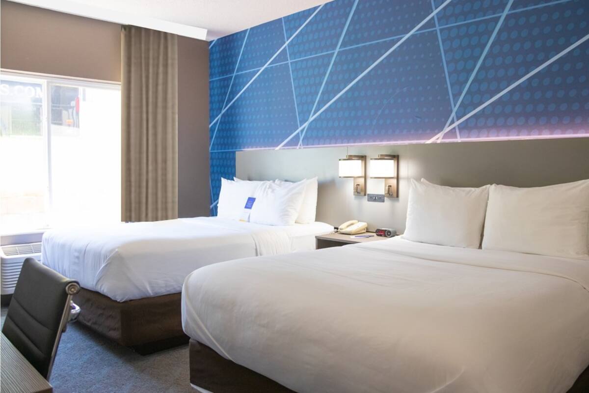 A hotel room with two beds, a desk, a chair, a nightstand with a phone, and a wall with a geometric pattern.