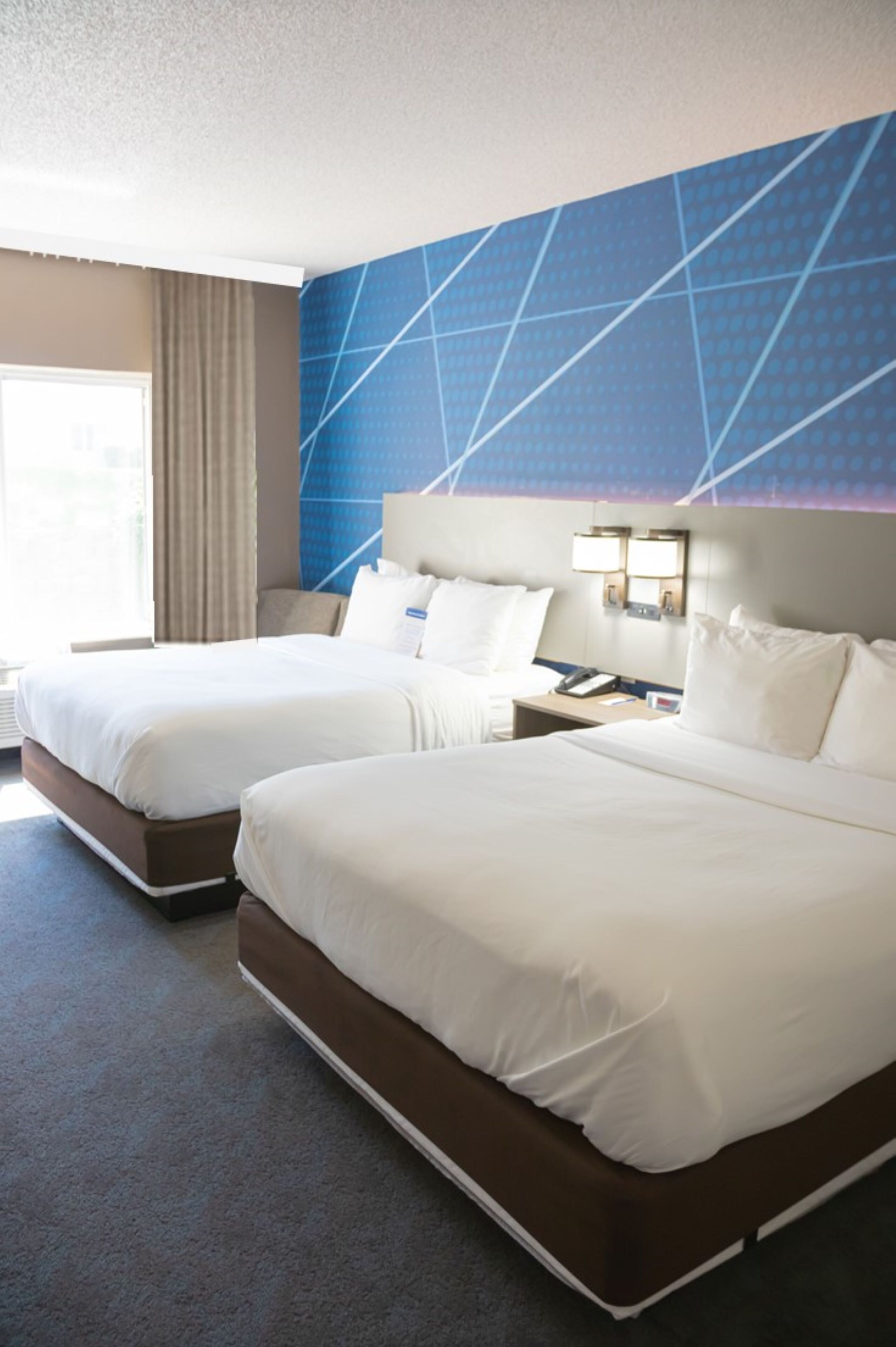 Habitación de hotel con dos camas, un ventanal con cortinas y un diseño mural azul geométrico.