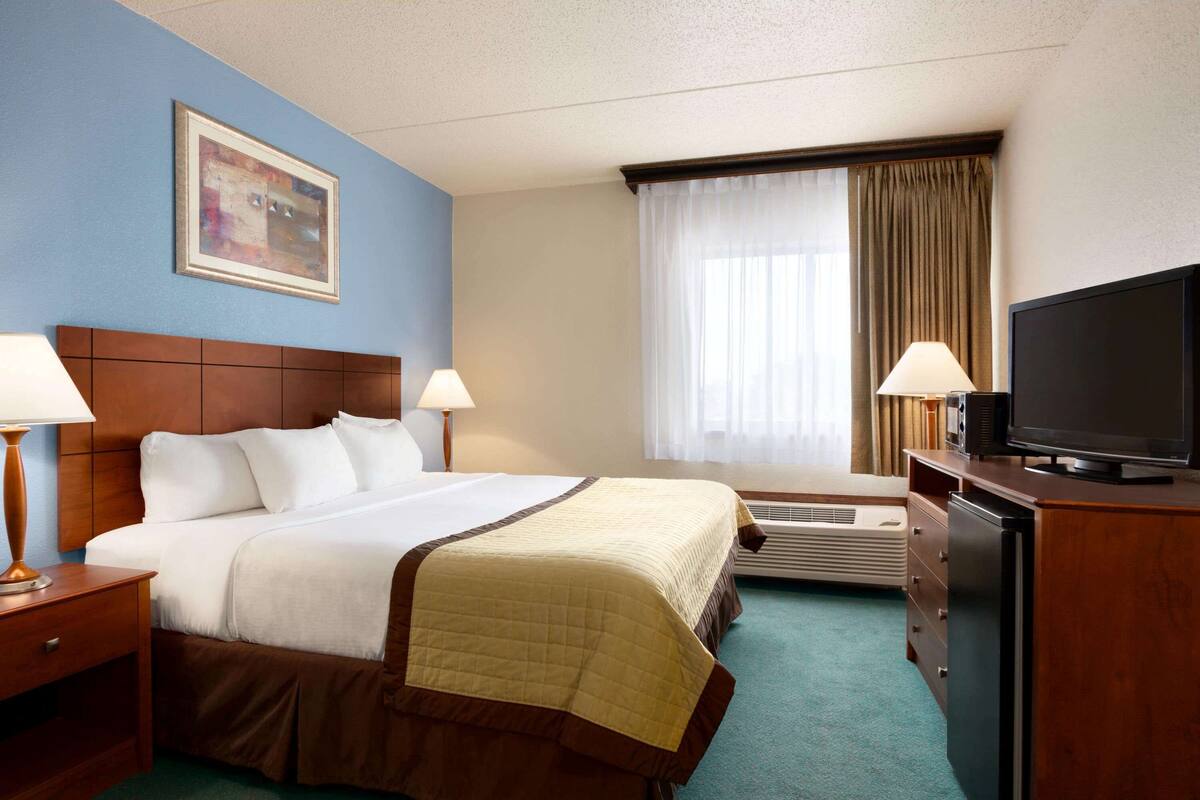 Une chambre d’hôtel avec un grand lit, deux tables de chevet avec des lampes, un téléviseur à écran plat, une commode et une fenêtre avec des rideaux.