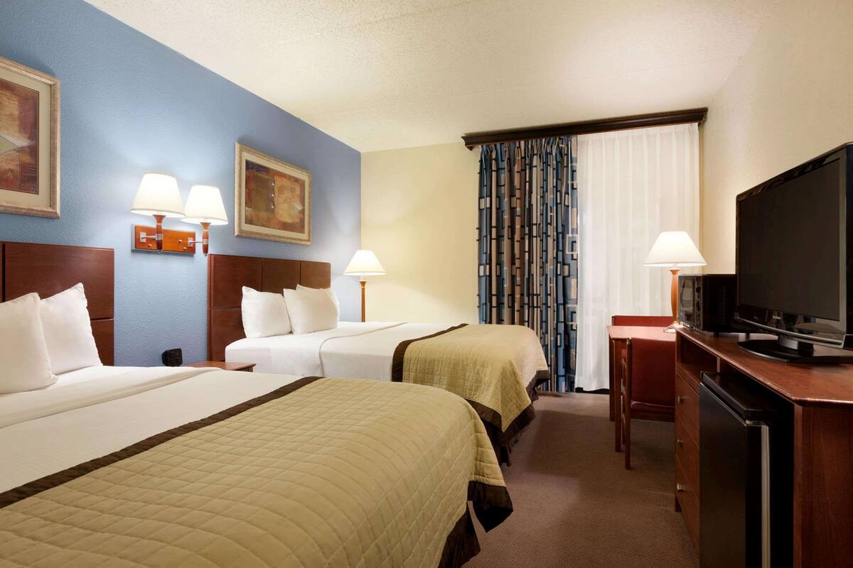 Une chambre d’hôtel avec deux lits, un bureau avec une télévision et une chaise. Il y a des lampes au mur et un tableau au-dessus des lits.