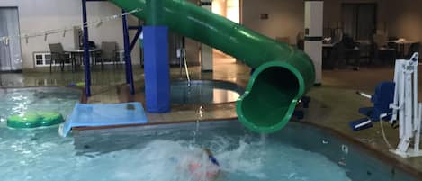 An indoor swimming pool with a green slide, a diving board, and a lifeguard stand.