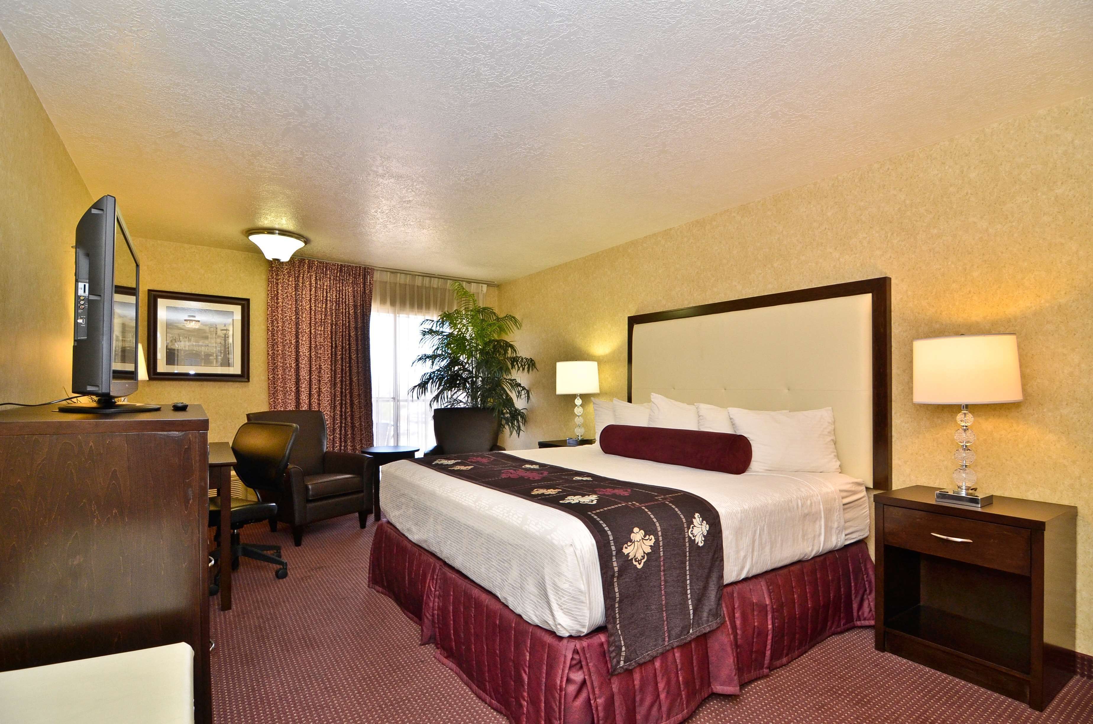 A hotel room with a large bed, a desk with a computer, a chair, a desk lamp, and a nightstand with a lamp.