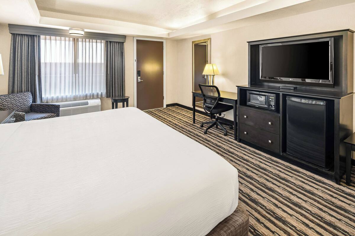 A hotel room with a large bed, a desk with a TV, a chair, a lamp, and a minibar.
