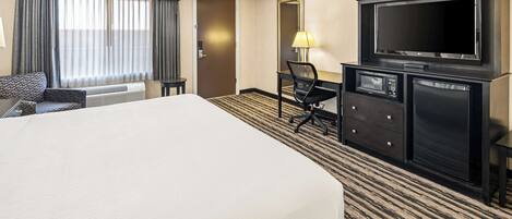 A hotel room with a large bed, a desk with a TV, a chair, a lamp, and a minibar.