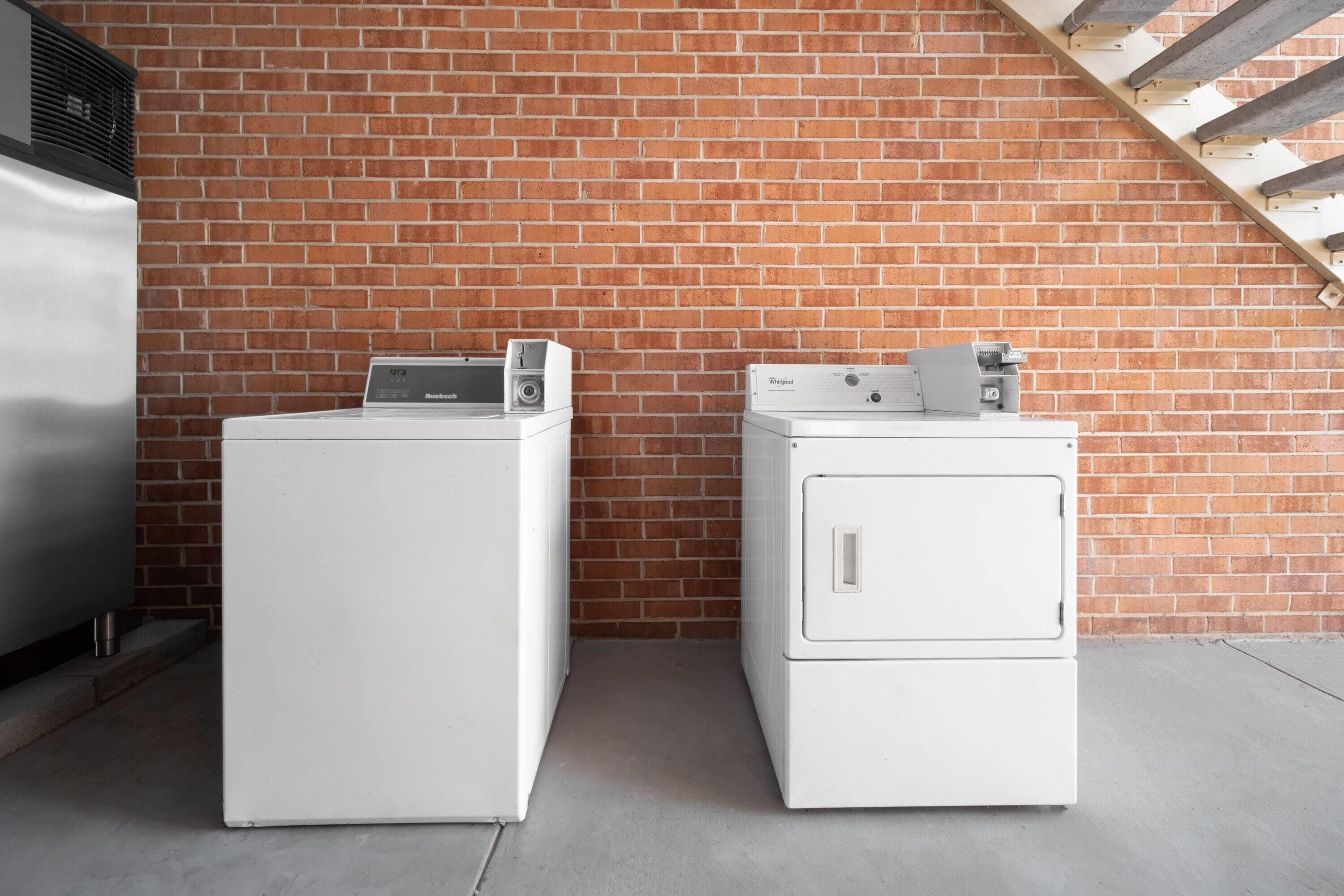 Deux appareils électroménagers blancs, une machine à laver et un sèche-linge, sont placés contre un mur de briques, et on aperçoit un escalier en bois à l’arrière-plan.