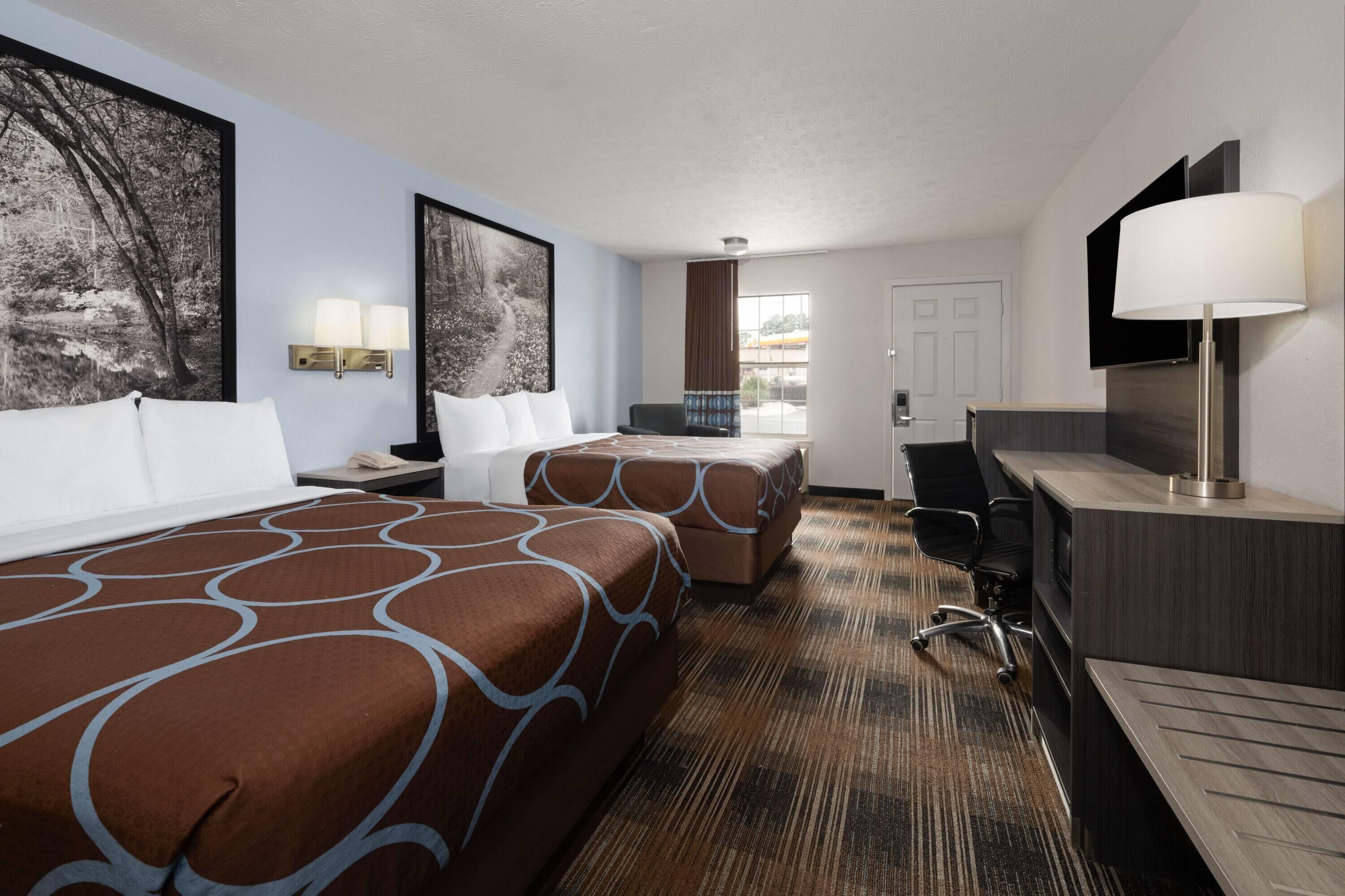 A hotel room with two beds, a desk, a chair, a TV, and artwork on the walls.
