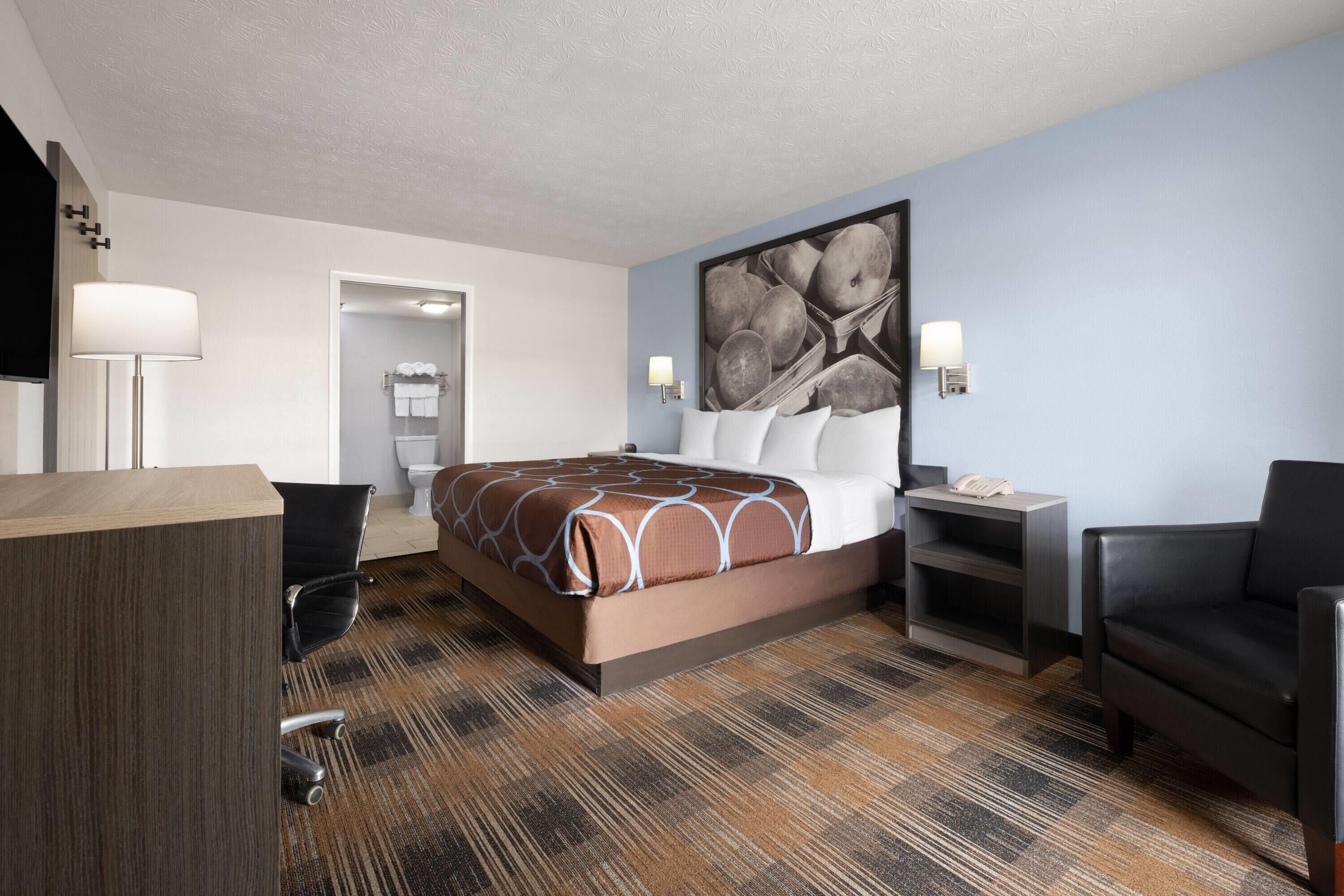 A hotel room with a bed, a desk, a chair, a lamp, and a painting on the wall.