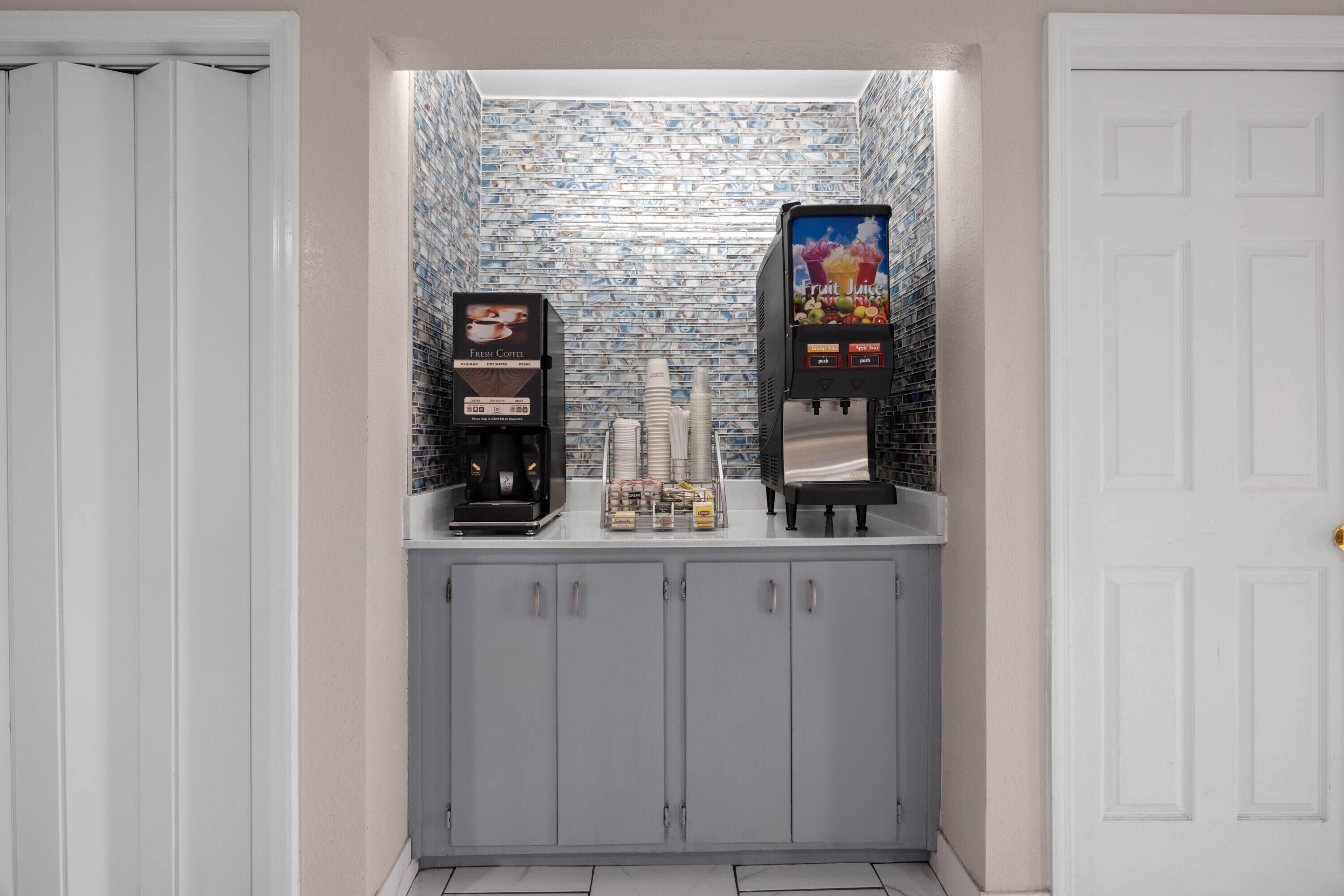A small kitchenette area with two coffee machines and a beverage dispenser, featuring a tiled backsplash and grey cabinets.