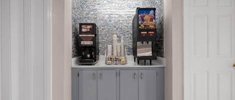 A small kitchenette area with two coffee machines and a beverage dispenser, featuring a tiled backsplash and grey cabinets.