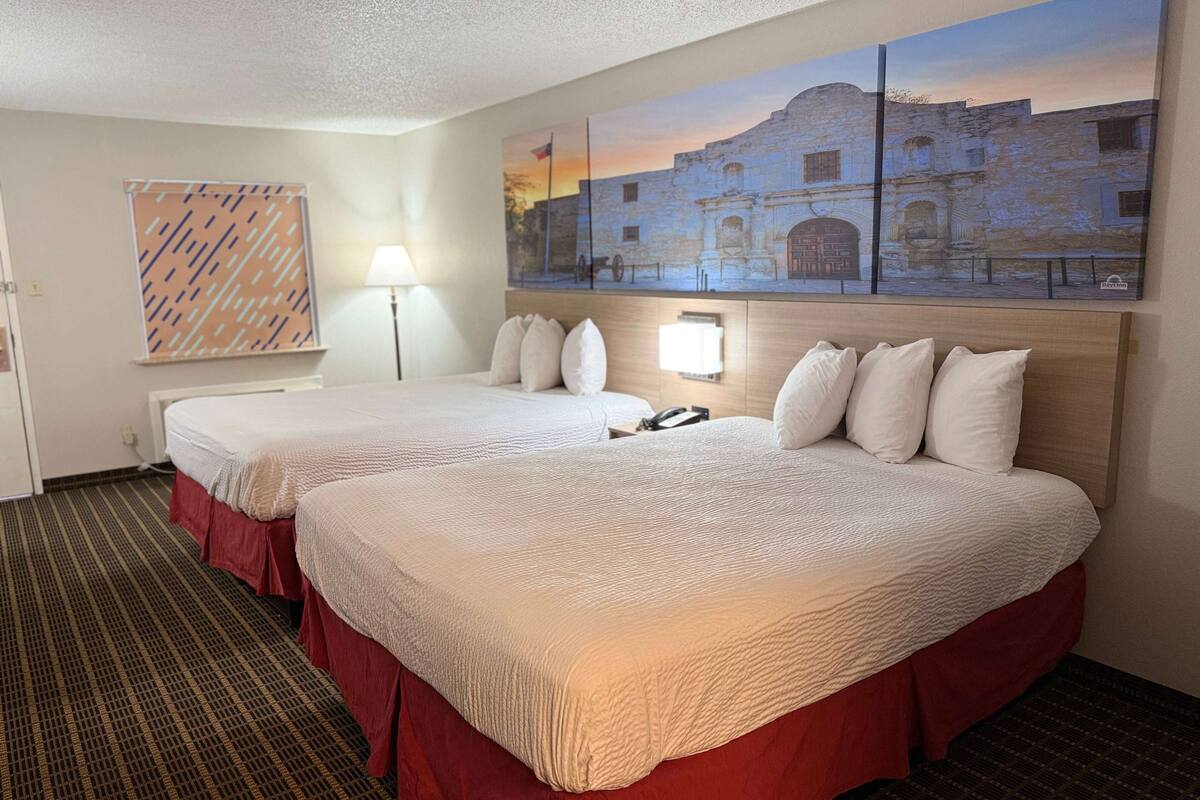 Quarto de hotel com duas camas, uma grande pintura de um edifício histórico, um painel de parede estampado e um abajur.