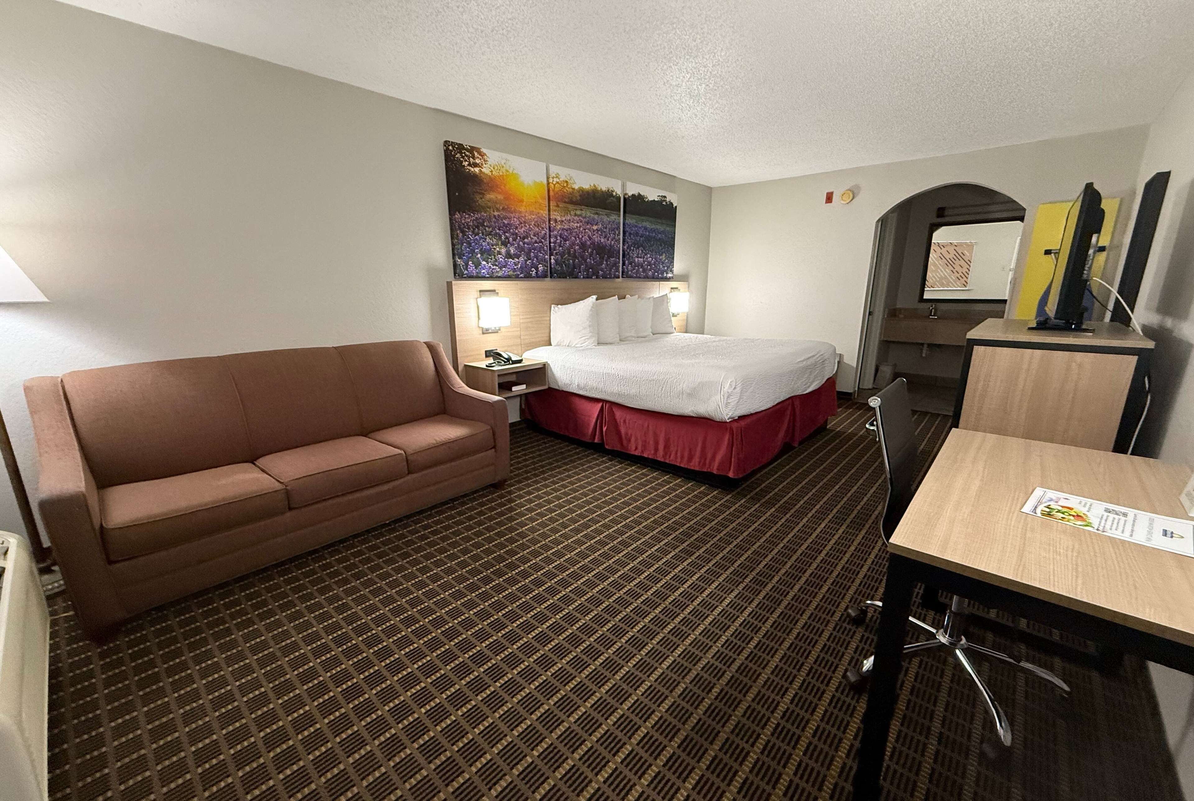 Una habitación de hotel con un sofá marrón, una cama con falda roja, un escritorio con una silla y un televisor de pared.