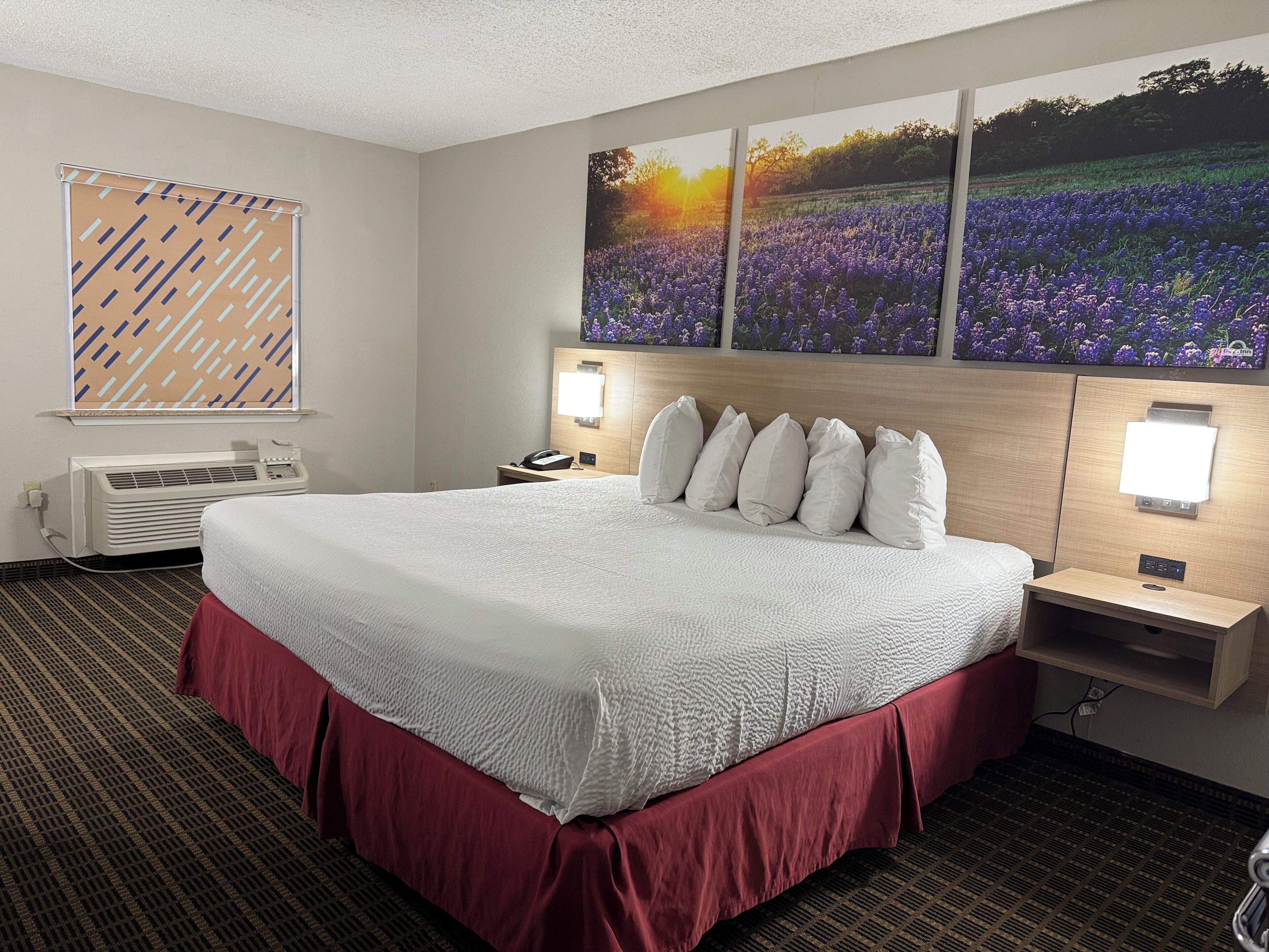 A hotel room with a large bed, a wall-mounted TV, a bedside table with a lamp, and a window with blinds.