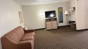 Habitación de hotel con un sofá marrón, un televisor de pantalla plana sobre un soporte de madera, un escritorio con lámpara y una alfombra estampada.