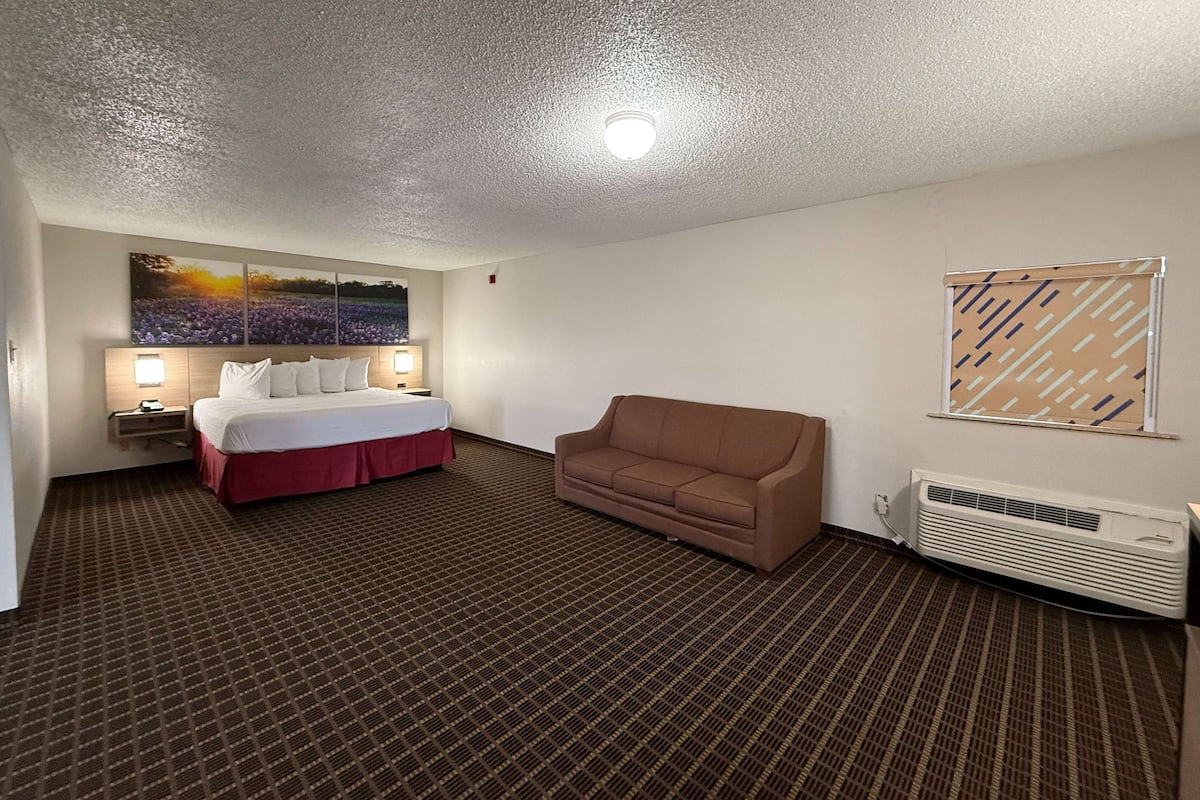 Quarto de hotel com uma cama, um sofá marrom, ar condicionado fixado na parede e uma grande pintura de paisagem.