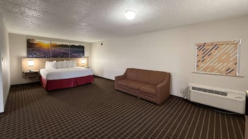 A hotel room with a bed, a brown sofa, a wall-mounted air conditioner, and a large landscape painting.