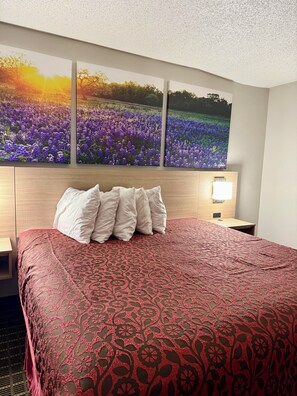 Habitación de hotel con una cama grande, cabecera con luz nocturna y tres cuadros de flores moradas colgados en la pared.