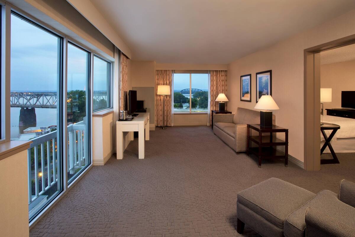A hotel room with a large window, a sofa, a table, and a TV.