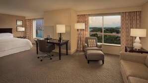 A hotel room with a large bed, a desk, two chairs, a sofa, and a view of a bridge and cityscape.