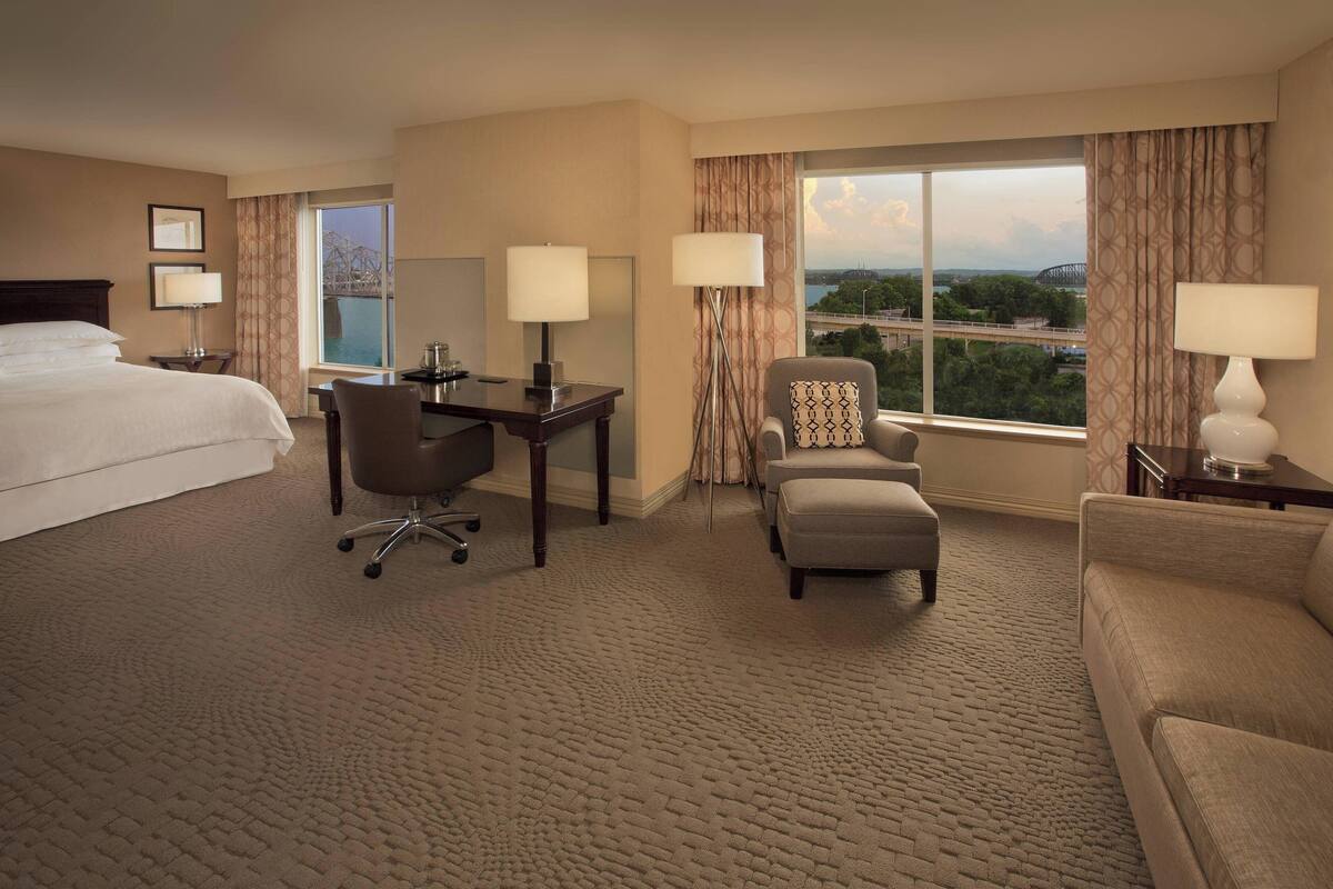 A hotel room with a large bed, a desk, two chairs, a sofa, and a view of a bridge and cityscape.