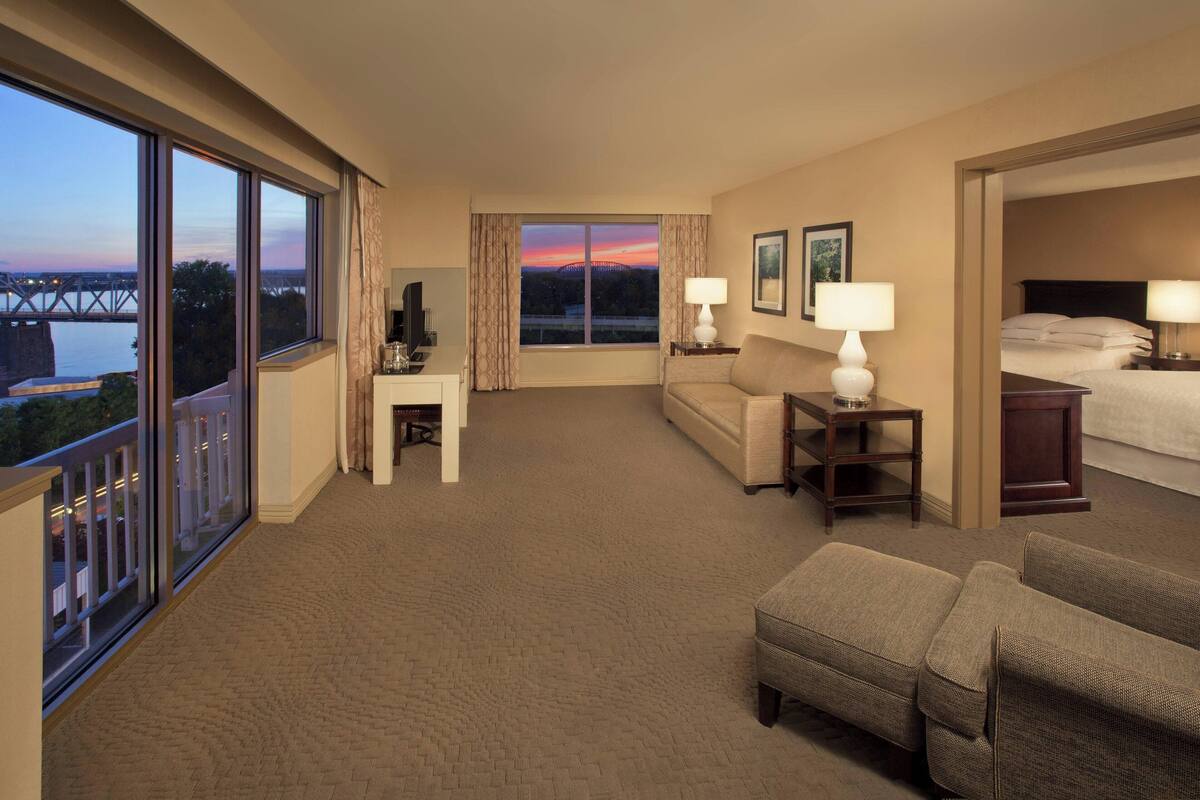 A hotel room with a large window, a sofa, a small table, a lamp, and a bed.