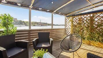 Um pátio ao ar livre com móveis de vime, uma mesa de vidro e vista para um rio e edifícios ao longe.