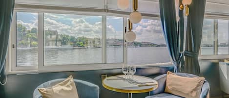 A dining area with two blue chairs, a round table, and a view of a river and buildings.