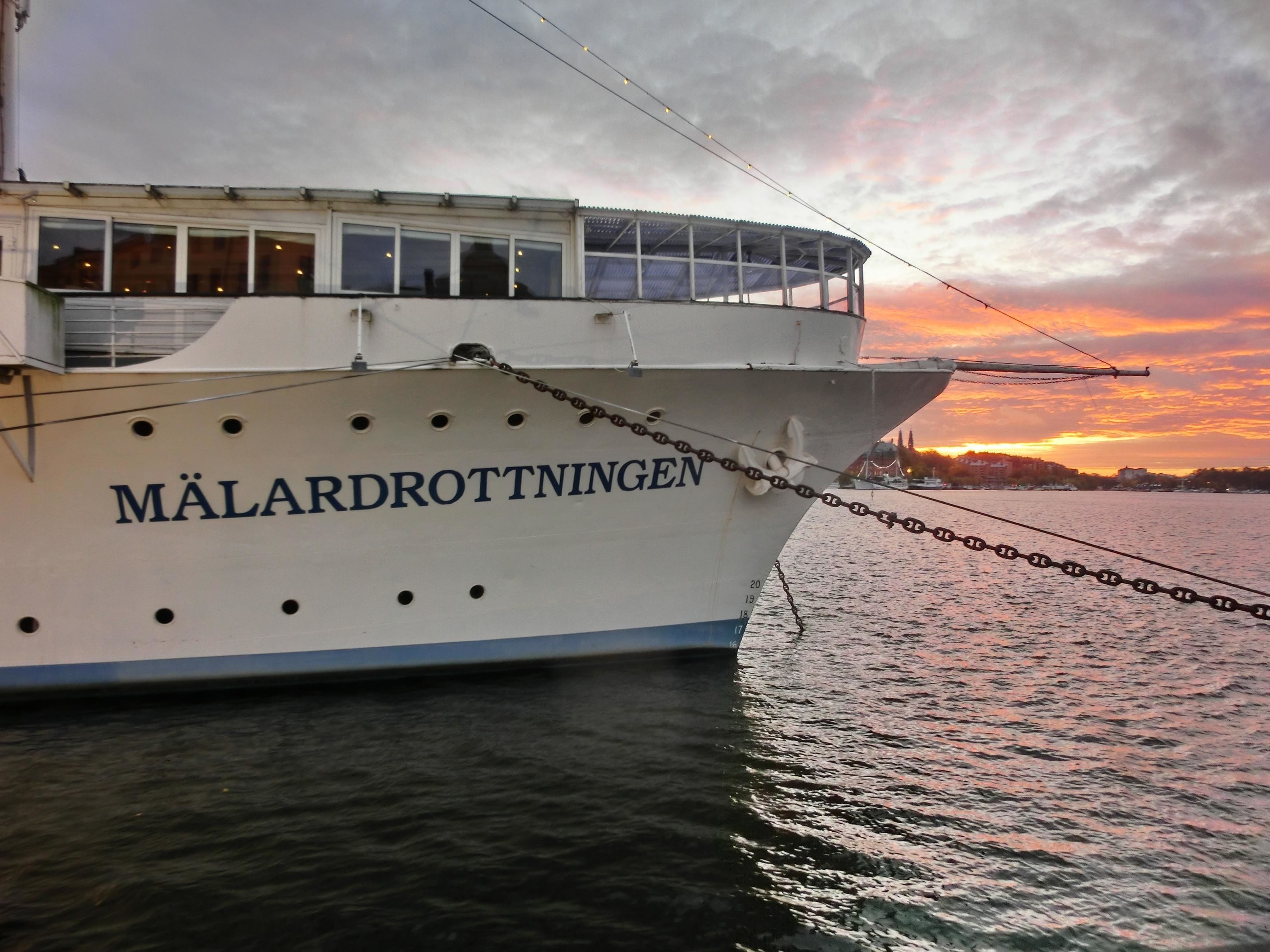 Un barco llamado "Målarodrottningen" atracado en un puerto con un atardecer de fondo.