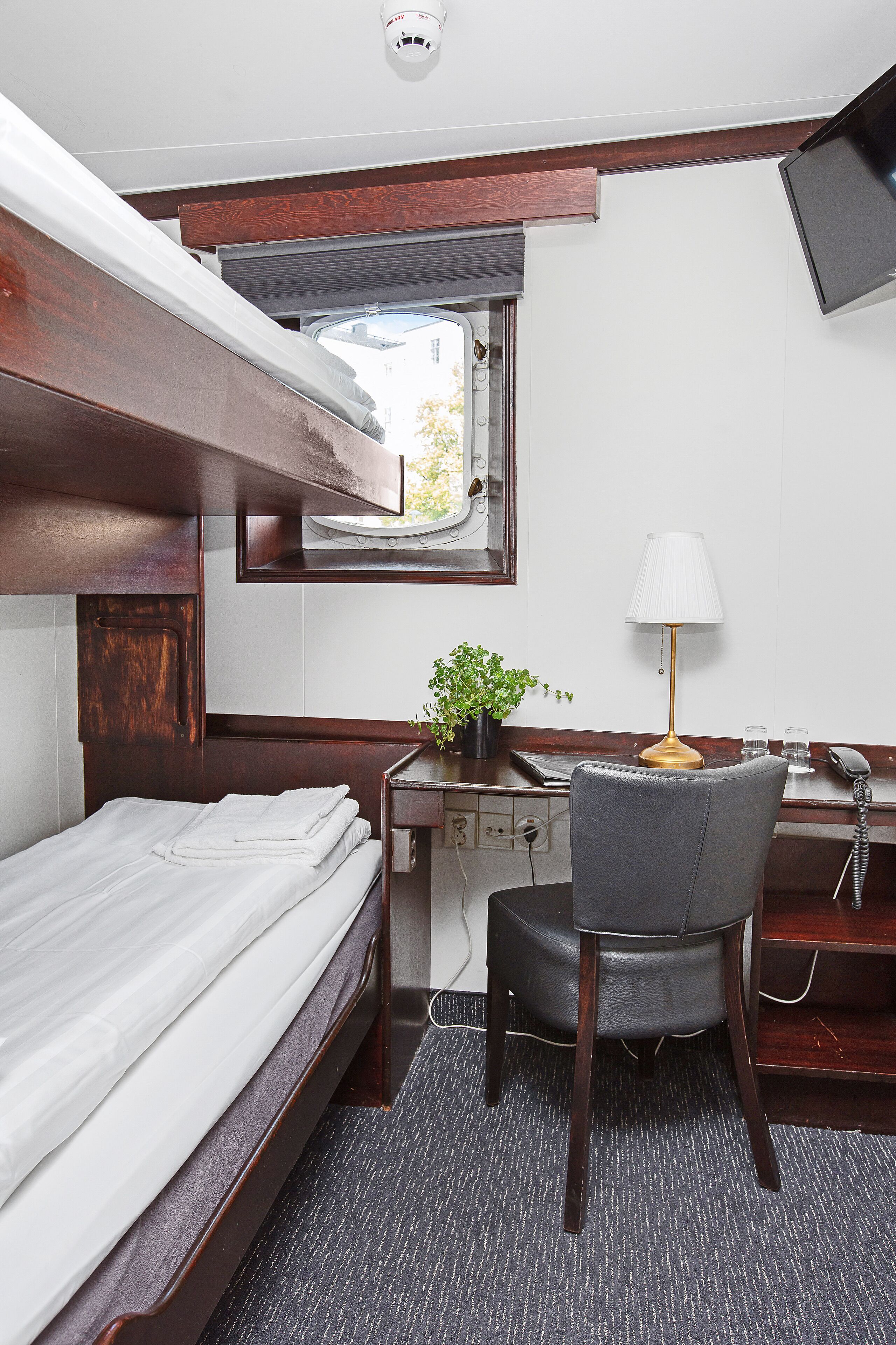 A compact hotel room with a bunk bed, a desk with a lamp, a chair, a small plant, and a window with a view.