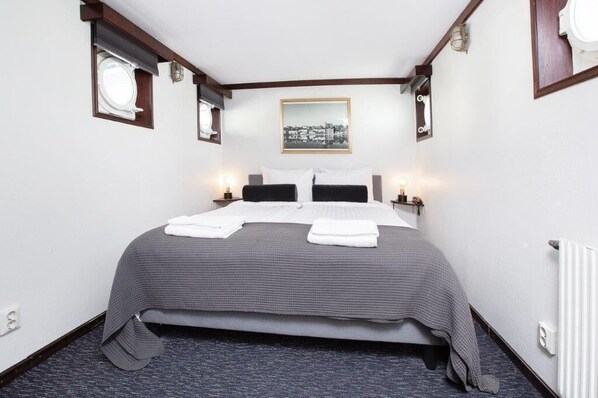 A neatly made bed with a grey bedspread and white pillows, a small window with a view, and a framed picture on the wall. - Mälardrottningen Hotel (Stockholm)