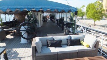 Una terraza exterior con zona de estar cubierta, una rueda grande y una fila de mesas y sillas.