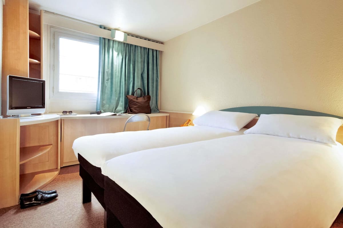 A hotel room with a bed, a desk with a computer monitor, a chair, a window with curtains, and a pair of shoes on the floor.