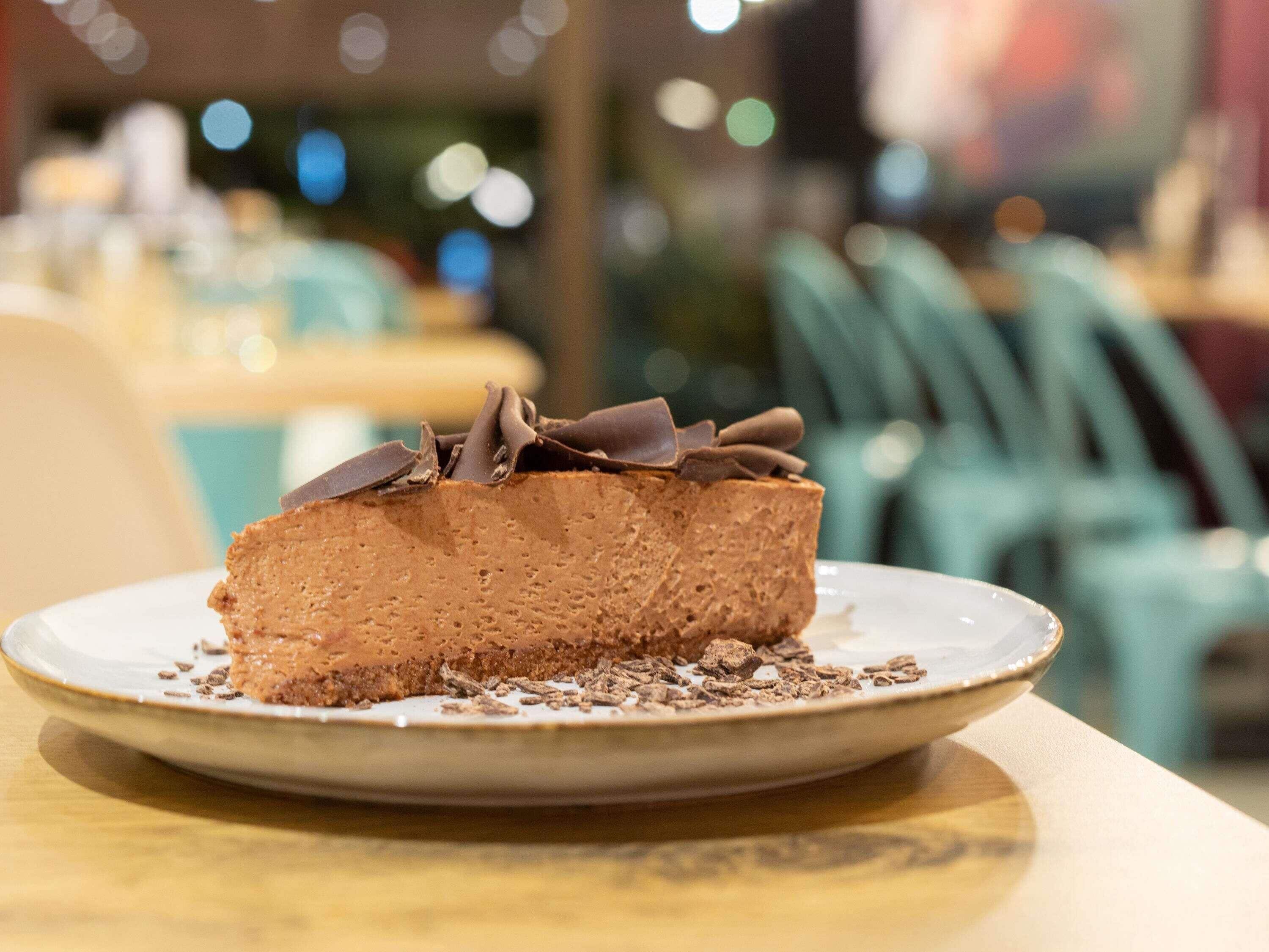 A slice of chocolate cake on a plate with chocolate shavings on top, placed on a table in a restaurant.
