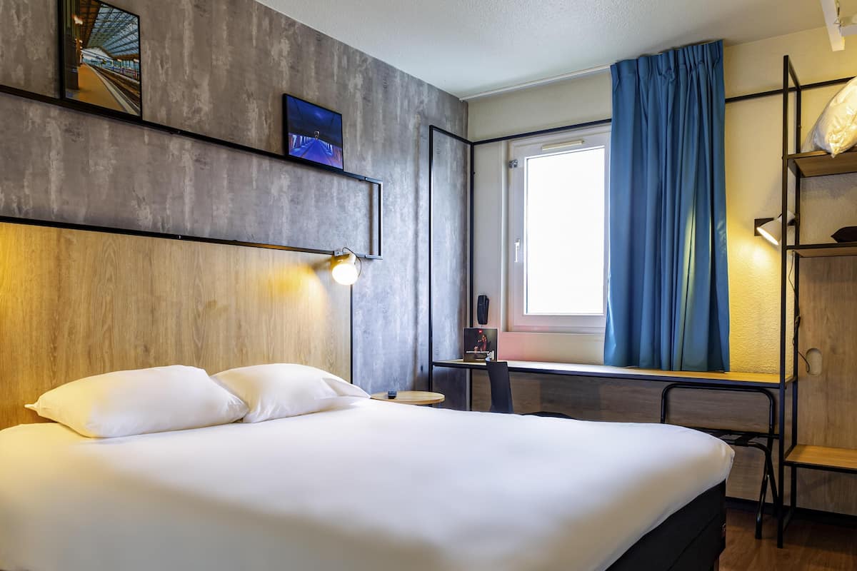 A modern hotel room with a large bed, a desk, a chair, a window with curtains, and a wall-mounted TV.