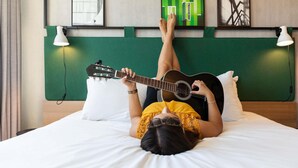 Una persona recostada en una cama con una guitarra, un cabecero verde y cuadros en la pared.