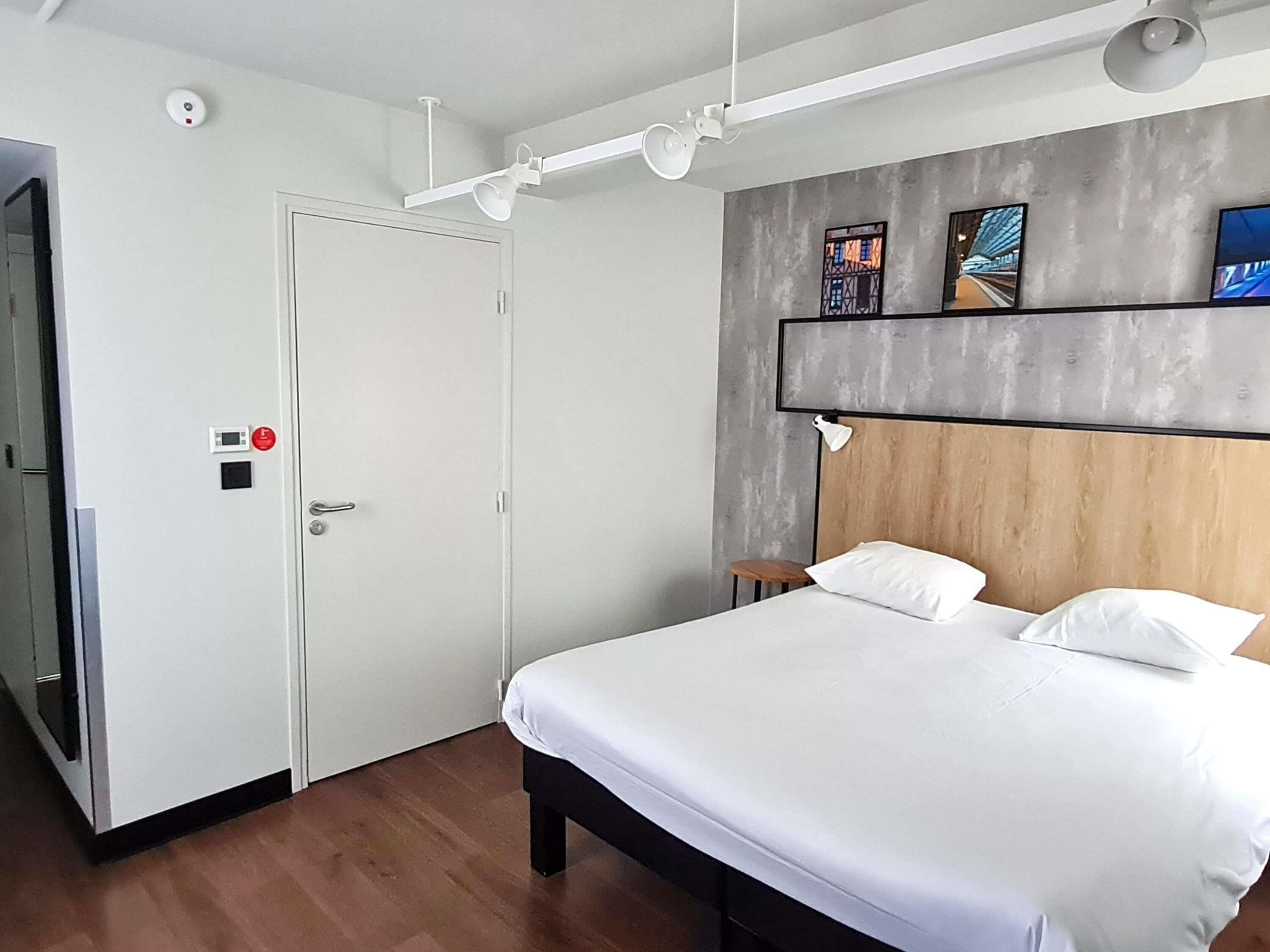 A modern hotel room with a bed, a white door, a small stool, and a wall-mounted shelf with pictures.