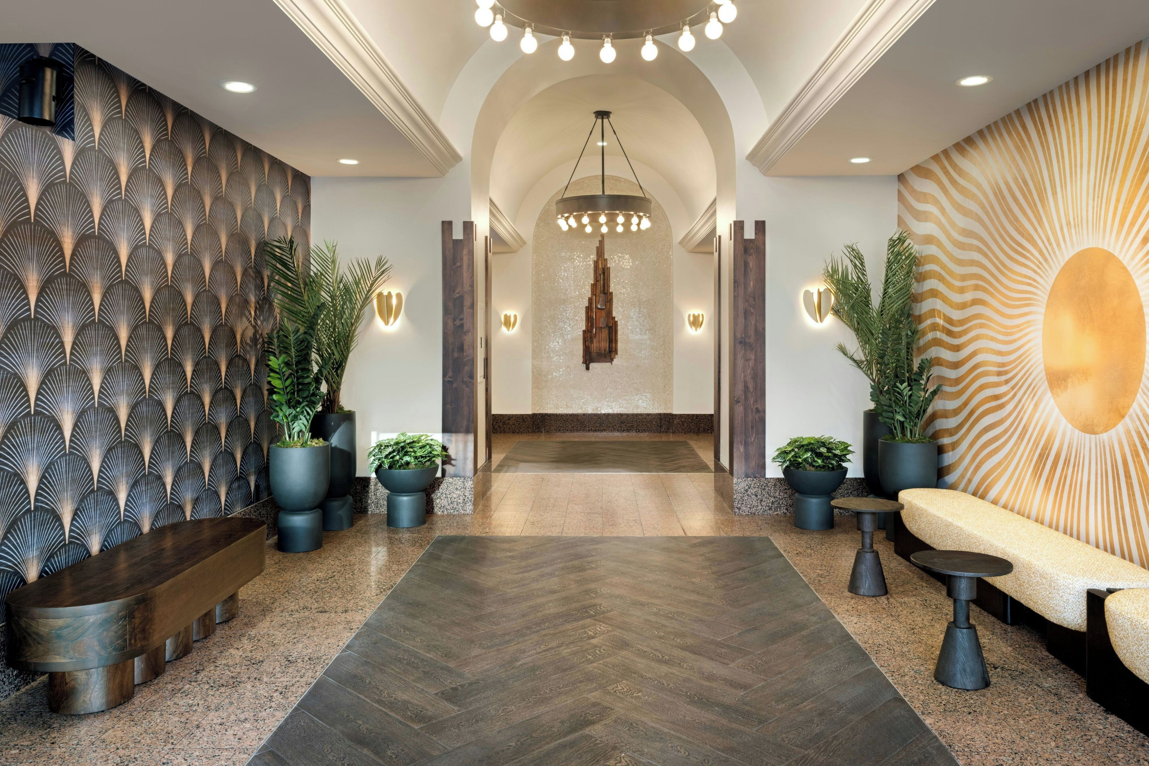 Un hall d’entrée moderne avec un mur orné d’un motif en chevrons, un lustre central et un espace salon agrémenté d’un décor en forme de soleil rayonnant.