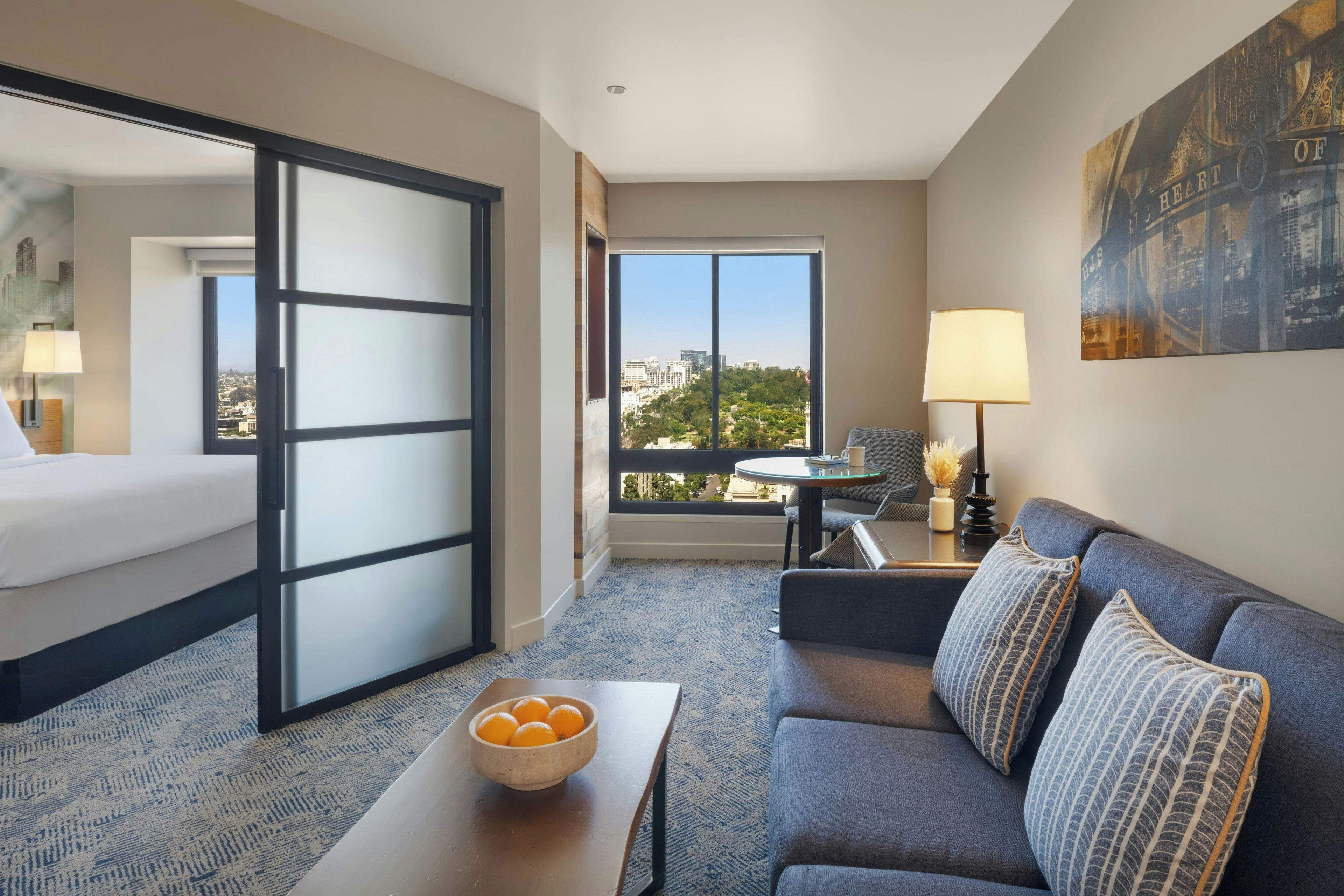 Habitación de hotel con cama, sofá, mesa con frutero, silla, lámpara y ventana grande con vista a la ciudad.