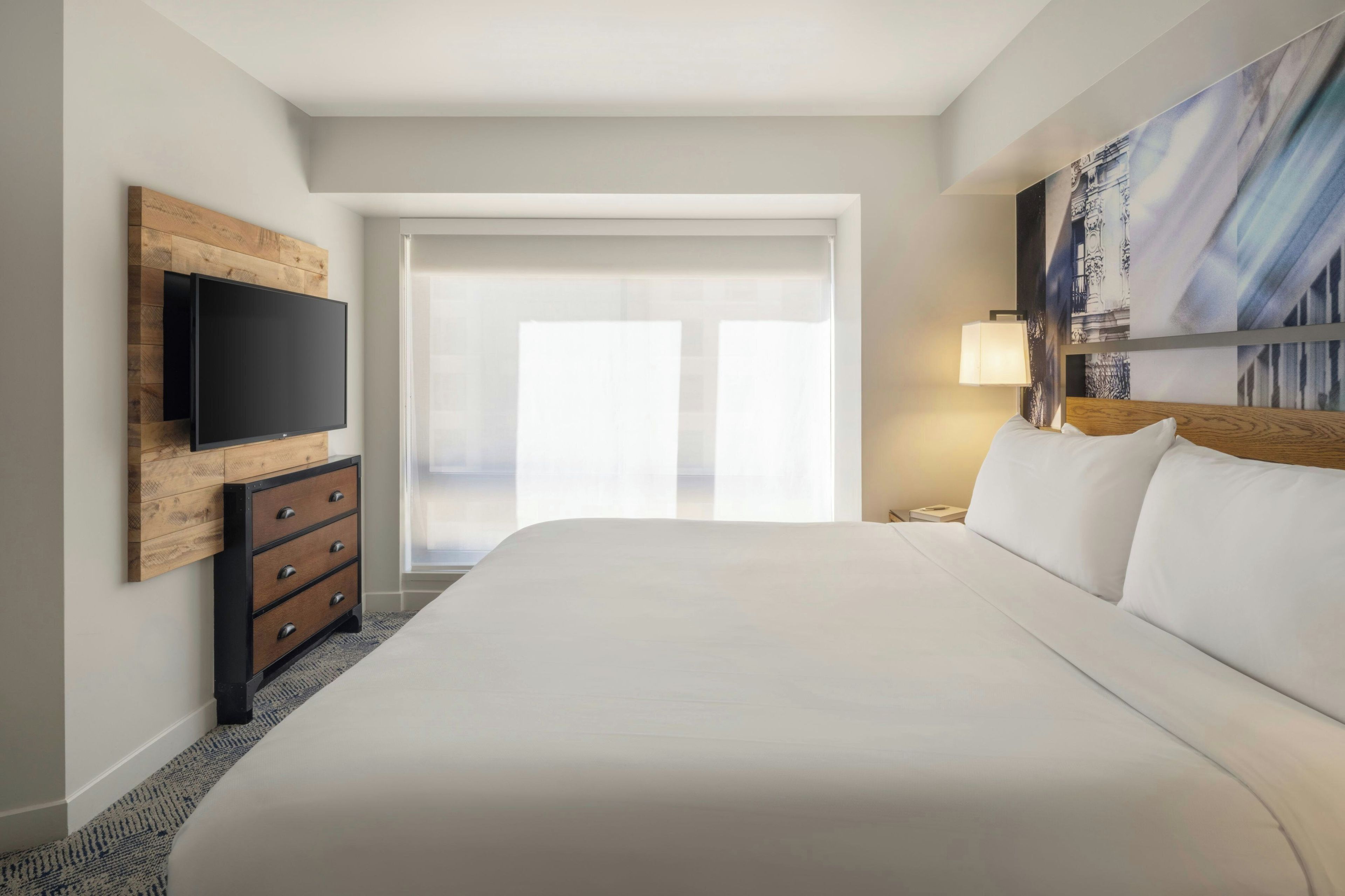 A hotel room with a large bed, a flat-screen TV mounted on a wooden panel, a nightstand with a lamp, and a large window with a view.