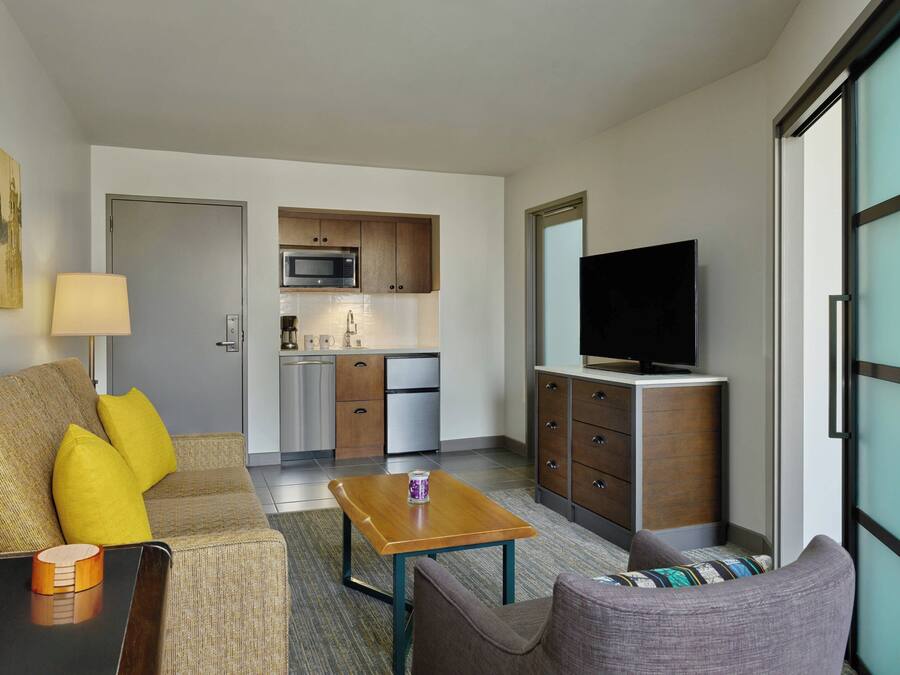 A compact hotel room with a kitchenette, a sofa with yellow cushions, a coffee table, a TV, and a sliding glass door.