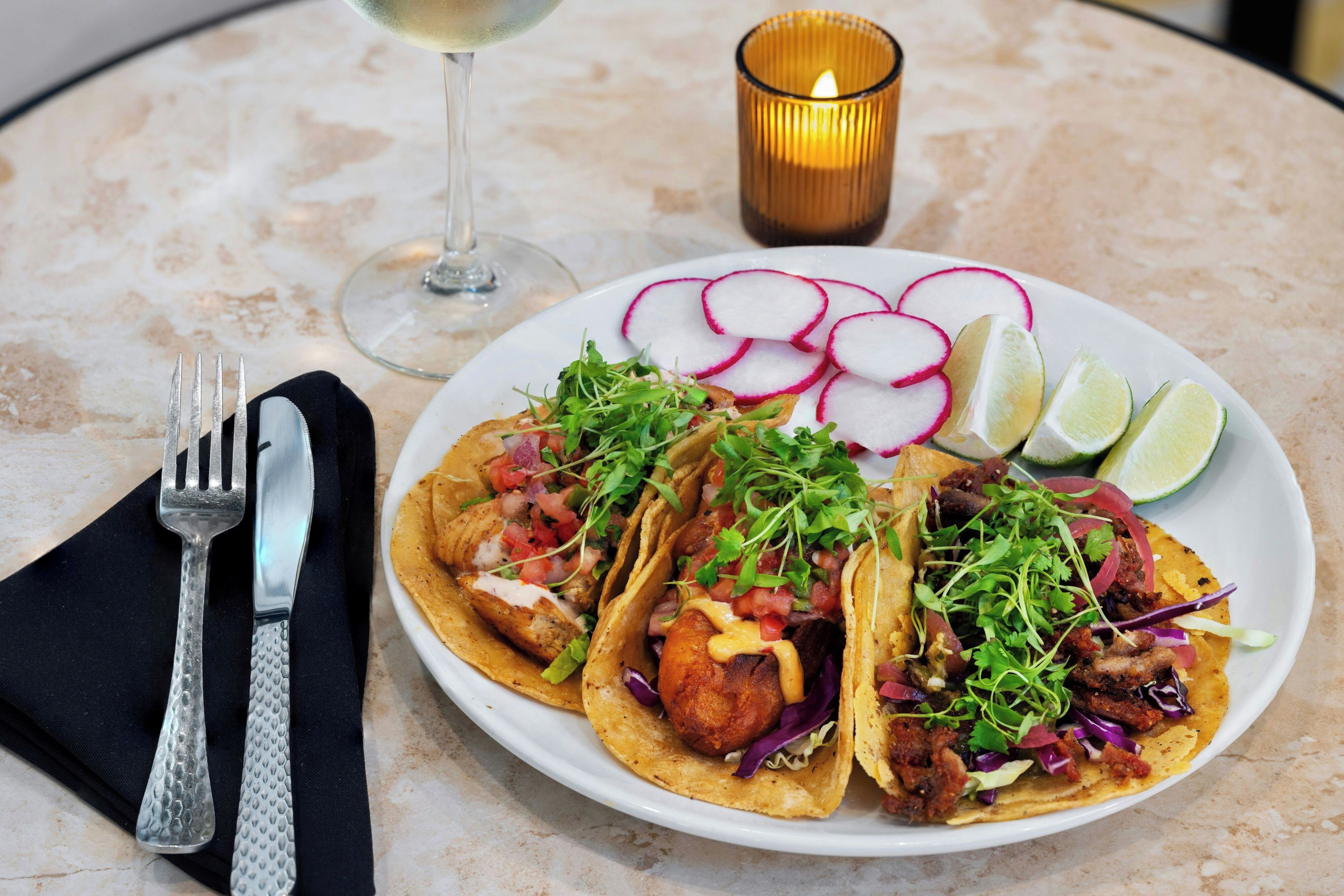 Ein Teller Tacos mit verschiedenen Belägen, ein Glas Wein und eine Kerze auf einem Tisch.