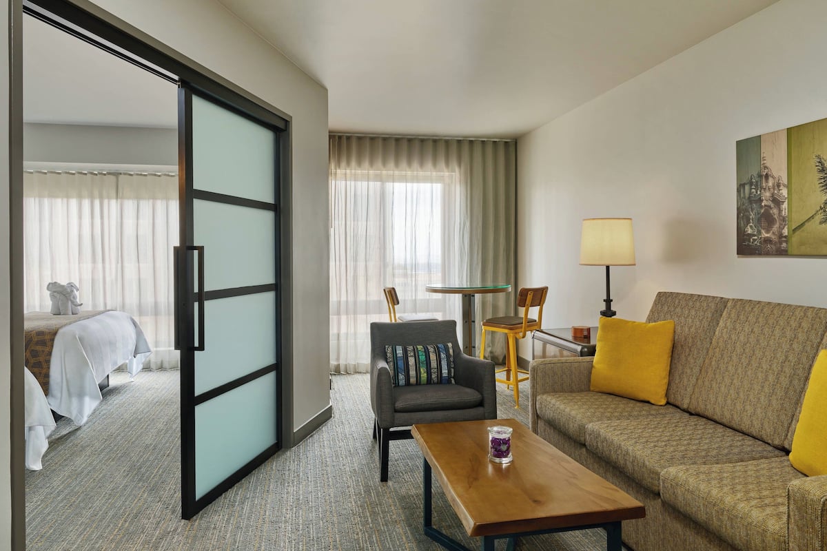 A hotel room with a bed, a sofa, a coffee table, a chair, a lamp, and a painting on the wall.