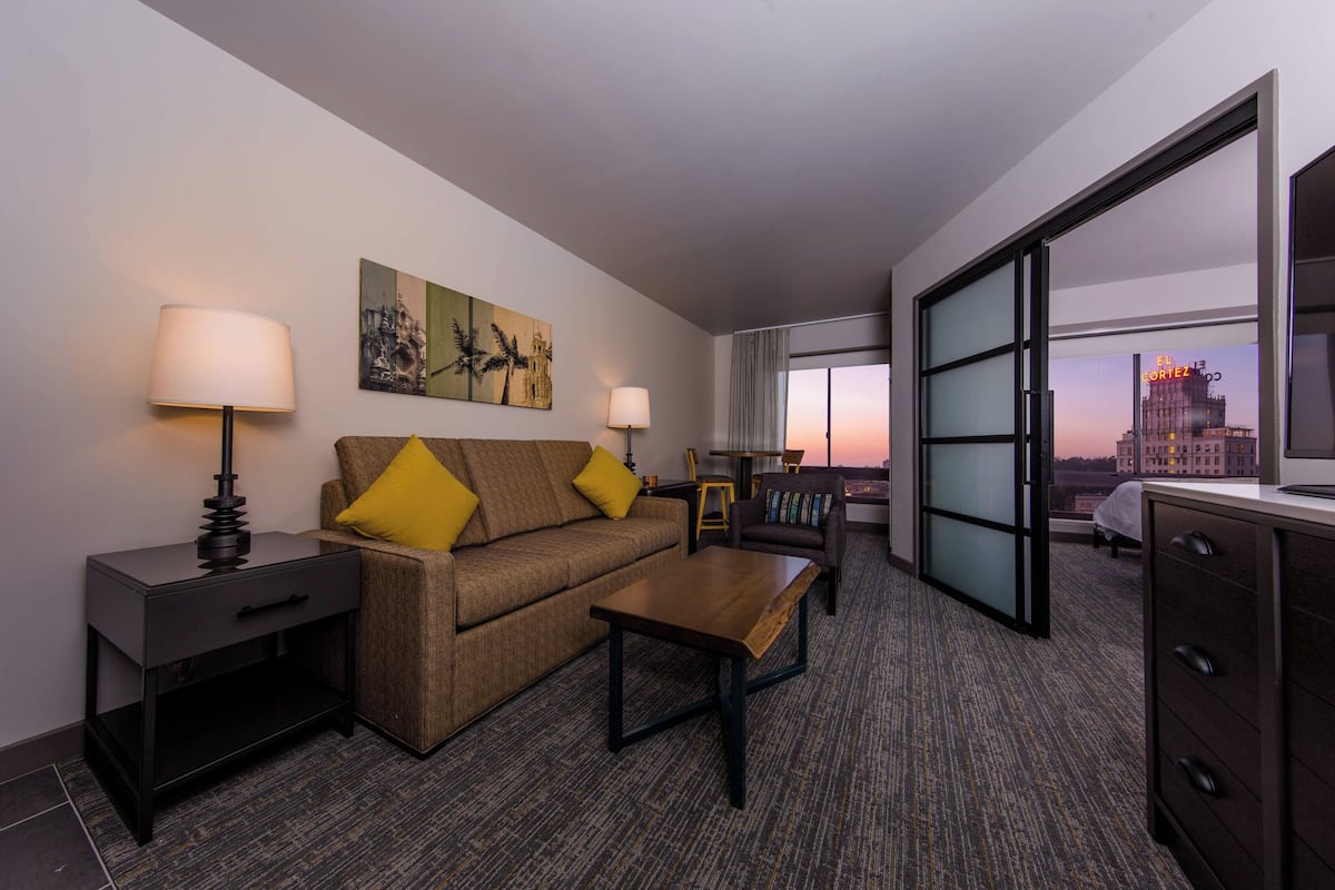 A hotel room with a sofa, two lamps, a coffee table, a desk, and a large window with a city view.