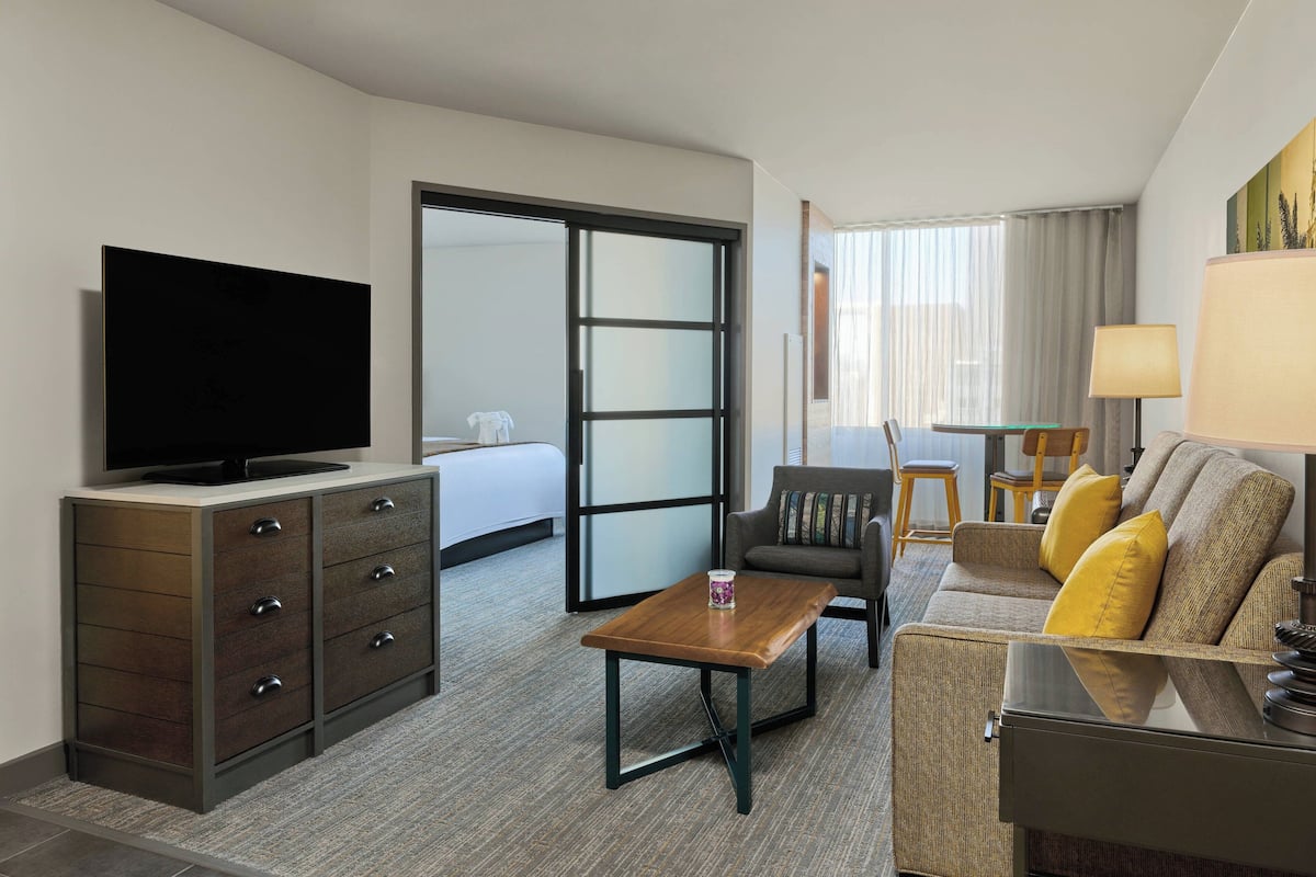 A hotel room with a television, a wooden desk, a sofa with cushions, a bed, and a sliding door leading to a balcony.