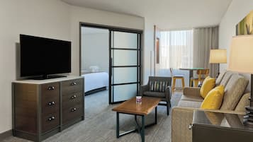 Quarto de hotel com televisão, secretária de madeira, sofá com almofadas, cama e porta deslizante para uma varanda.